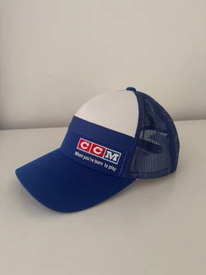 Blå och vit truckerkeps från CCM - Snygg blå och vit truckerkeps från CCM med broderad logga och texten 'When you're born to play' framtill. Kepsen har mesh på sidorna och bak för extra ventilation samt böjd skärm. Perfekt för dig som gillar sportig stil.