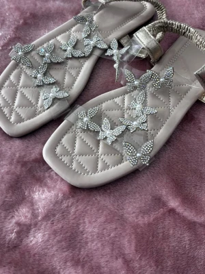 Beige sandaler med strassfjärilar - Säljer ett par beige sandaler med quiltad sula och fyrkantig tå. Dekorerade med glittrande strassfjärilar och transparenta band. Elastisk rem bak för skön passform. Perfekta för dig som gillar detaljer och vill sticka ut. använda 1 gångoch super fina.