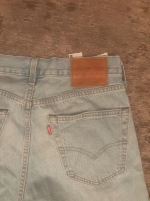 Levi's 501 ljusblå raka jeans - Klassiska Levi's 501 jeans i ljusblå denim med raka ben och hög midja. Jeansen har två bakfickor, den ikoniska röda Levi's-lappen och läderpatch bak i midjan. Perfekta för en avslappnad och trendig look.