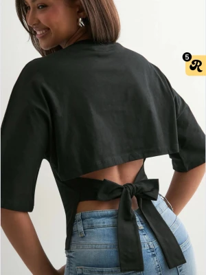 Populära Nelly toppen💖💖 - Trendig svart topp med halvlånga ärmar och en unik öppen ryggdetalj som knyts med en stor rosett baktill. Toppen har en croppad passform och är tillverkad i mjuk bomull, perfekt för en cool och avslappnad stil.