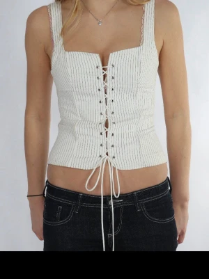 Vit korsettopp med snörning - Säljer en vit korsettopp med tunna svarta ränder och snörning framtill. Toppen har breda axelband, croppad passform och dragkedja i ryggen. Materialet känns som bomull eller polyester och toppen har en snygg, figurnära siluett.