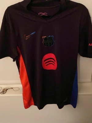 FC Barcelona Nike matchtröja svart - Snygg svart FC Barcelona matchtröja från Nike med Dri-FIT-material. Tröjan har klubbmärke, Nike-logga och Spotify-logga i rött framtill samt röda och blå detaljer på sidorna. Perfekt för fotbollsträning eller att bära som supporter.