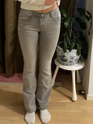 Grå bootcut jeans med låg midja - Säljer ett par gråa bootcut jeans med låg midja och klassiska fem fickor. Jeansen har en snygg tvättad look och är tillverkade i ett stretchigt denimtyg som sitter tajt över låren och släpper ut vid vaden. Perfekta för en avslappnad och trendig stil.