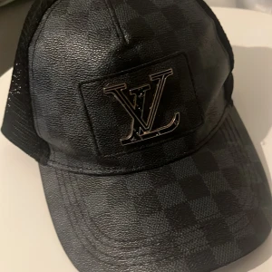 Svart Louis Vuitton keps i läderlook - Snygg svart keps från Louis Vuitton med klassiskt rutmönster och LV-logga i metall framtill. Kepsen har mesh på baksidan för extra ventilation och en stilren läderlook som ger en exklusiv känsla. Perfekt accessoar för dig som gillar streetstyle och lyx.