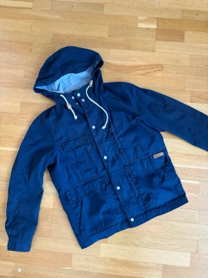 Mörkblå vindjacka med huva - Snygg mörkblå vindjacka från Didriksons med justerbar huva och vita dragsnören. Jackan har fyra stora fickor framtill, tryckknappar och dragkedja. Materialet är slitstarkt och passar perfekt för blåsiga dagar.