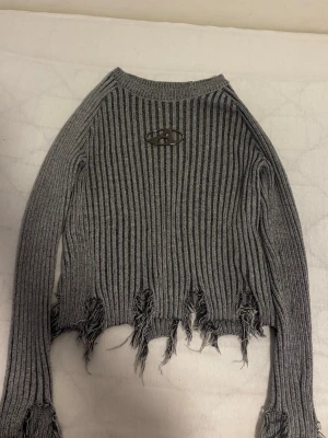 Grå ribbstickad tröja med slitningar - Trendig grå ribbstickad tröja med råa, slitna kanter och ett metallfärgat emblem på bröstet. Långärmad modell med croppad passform och markerade muddar. Perfekt för en edgy streetwear-look.