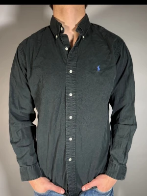 Svart skjorta från Ralph Lauren - Klassisk svart skjorta från Ralph Lauren med button-down krage och blå broderad logga på bröstet. Skjortan har långa ärmar, vita knappar och är tillverkad i bomull. Perfekt för en stilren och avslappnad look.