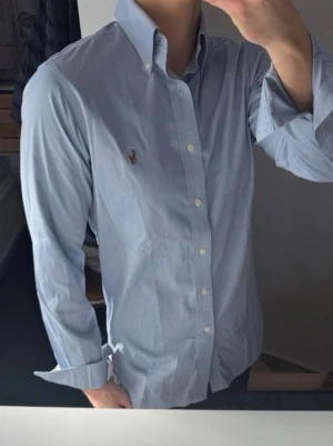 Ljusblå skjorta från Ralph Lauren - Klassisk ljusblå skjorta från Ralph Lauren med button-down krage och broderad logga på bröstet. Skjortan har långa ärmar, vita knappar och är tillverkad i mjuk bomull. Perfekt för en clean och stilren look.