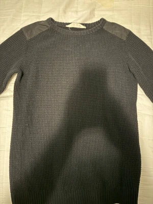 Mörkgrå stickad tröja från H&M - Snygg mörkgrå stickad tröja från H&M med förstärkta axelpartier i grått. Tröjan har lång ärm och en grovstickad struktur som ger en cool och avslappnad look. Perfekt till jeans eller chinos.