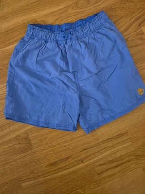 Blå shorts från Timberland - Snygga blå shorts från Timberland med elastisk midja och sidofickor. Liten gul Timberland-logga broderad på vänster ben. Perfekta för varma dagar och enkel att matcha med olika toppar.