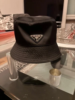 Svart bucket hat från Prada - Snygg svart bucket hat från Prada i glansigt nylonmaterial. Klassisk form med bred, nedåtlutande brätte och ikonisk metalltriangel med logga framtill. Perfekt accessoar för en trendig och stilren look.