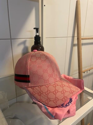 Rosa Gucci keps - Säljer en rosa keps från Gucci med klassiskt GG-monogram över hela kepsen och en svart-röd rand på sidan. Kepsen har justerbart band bak och är tillverkad i bomullsmix. Perfekt för dig som vill sticka ut med en lyxig accessoar.