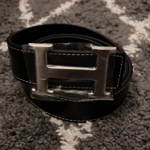 H-bälte  - Snyggt bälte i svart och med  svart skinn med ikoniskt,H-spänne i silverfärgad metall. Bältet har stilrena, kontrastsömmar och klassisk design som lyfter, vilken outfit som helst. Strl 110cm