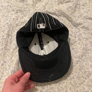 Svart LA-keps med vita ränder - Snygg svart keps från New Era med vita vertikala ränder och LA-logga framtill. Klassisk böjd skärm med vita sömmar och broderad MLB-logga på baksidan. Justerbar passform och tillverkad i bomullsmaterial. Perfekt för dig som gillar streetwear och sportig stil.