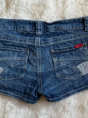Blå jeansshorts från ONLY - Säljer ett par klassiska blå jeansshorts från ONLY med fem fickor, vita kontrastsömmar och uppvikta benslut. Shortsen har låg midja och en snygg tvättad denimlook. Perfekta för varma dagar och enkel att matcha med allt. 