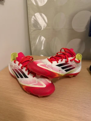 Adidas X Speedflow fotbollsskor - Säljer ett par Adidas X Speedflow fotbollsskor med snygg design i vitt, rött och gult. Skorna har röda snören, svarta och röda detaljer på sidan samt gula inslag vid hälen. Tillverkade i lätt syntetmaterial för maxad fart på planen.