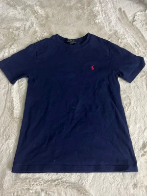 Mörkblå t-shirt från Polo Ralph Lauren - Klassisk mörkblå t-shirt från Polo Ralph Lauren med röd broderad logga på bröstet. T-shirten har rund halsringning och korta ärmar. Tillverkad i mjuk bomull för en skön känsla och enkel stil.