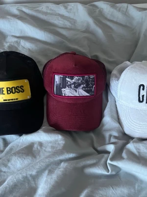 Vinröd truckerkeps med tryck - Säljer en vinröd truckerkeps med mesh på baksidan och justerbar snapback. Framtill finns ett svartvitt tryck med motiv av personer och texten 'KNS S'. Perfekt för dig som vill ha en unik och stilren keps med streetkänsla. Säljer 5 kepsar för 350kr man ser att dom är använts men de var ett tag sen jag använde dom 