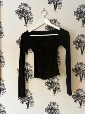 Svart långärmad topp med rosett - Trendig svart långärmad topp med liten rosett framtill och lätt rynkad detalj vid bysten. Toppen har en croppad passform och vågig kant nertill. Perfekt för dig som gillar stilrena plagg med en söt twist.