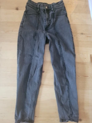 Grå jeans med hög midja - Säljer ett par grå jeans med hög midja och raka ben. Jeansen har tydliga sömmar framtill och klassisk femficksdesign. Passar dig som gillar en enkel och stilren look. Materialet är denim.