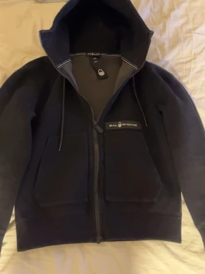 Marinblå hoodie från Sail Racing - Snygg marinblå hoodie från Sail Racing med dragkedja framtill, två stora fickor och justerbar huva med snören. Logotypen sitter på bröstet och insidan har en mjuk finish. Perfekt för en avslappnad och sportig stil.