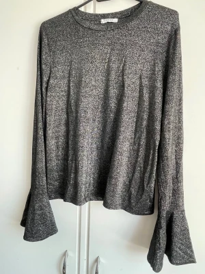 Glittrig svart topp med trumpetärm - Snygg svart topp från Pieces med glittrigt tyg och långa trumpetärmar. Perfekt för dig som vill sticka ut lite extra. Tunn och mjuk kvalitet med rund halsringning och relaxed fit. Aldrig använt faktiskt bara legat i garderoben utan att ha kommit till användning, Perfekt till nyår😻