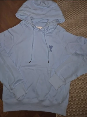 Ljusblå hoodie från Ami Paris ingår original påse - Ljusblå hoodie från Ami Paris med klassisk huva och dragsko. På bröstet finns det ikoniska hjärta-A-loggan i mörkblått. Hoodien har känguruficka och ribbade muddar vid ärmslut och nederkant. Perfekt för en chill och stilren look.
