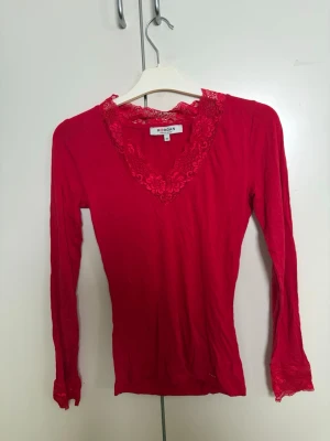 Röd långärmad topp med spets Morgan - Snygg röd långärmad topp från Morgan med v-ringning och spetsdetaljer vid halsringning och ärmslut. Figurnära passform och mjukt material som känns bekvämt mot huden. Perfekt för dig som vill ha en feminin och trendig look.