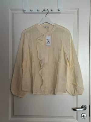 Krämvit blus med volang från Cubus - Stilren krämvit blus från Cubus med rund halsringning och dekorativ volang längs knäppningen framtill. Blusen har långa, lätt puffiga ärmar med resår vid ärmslut. Tillverkad i ett tunt, lätt glansigt material som ger en elegant känsla.