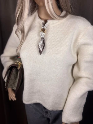 Vit stickad tröja med scarfdetalj - Snygg vit stickad tröja med rund hals och unik scarfdetalj i halsen med guldspänne. Tröjan har en rak passform och långa ärmar, perfekt för en trendig och stilren look. Passar till både jeans och kjol för en cool vardagsstil.