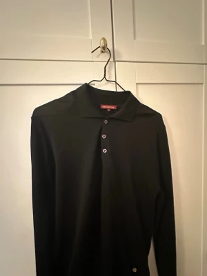 Svart långärmad pikétröja Nikolaj d’Étoiles - Stilren svart långärmad pikétröja från Nikolaj d’Étoiles med klassisk krage och tre knappar framtill. Tröjan är tillverkad i merinoull och har en mycket fin kvalitet. Är bara använd ett fåtal gånger pga för liten för mig. 