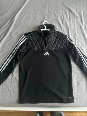 Svart träningsjacka från Adidas - Svart långärmad träningsjacka från Adidas med vita ränder längs ärmarna och quiltad detalj över bröstet. Halv dragkedja framtill och hög krage. Tillverkad i funktionsmaterial med Aeroready-teknologi för bra ventilation.