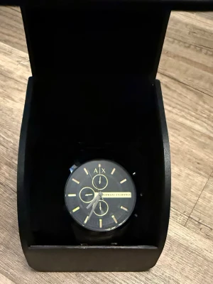 Svart klocka från Armani Exchange - Stilren svart klocka från Armani Exchange med justerbart stålarmband och guldfärgade detaljer på urtavlan. Klockan har kronograf-funktion och tydliga indexmarkeringar. Perfekt accessoar för dig som gillar en modern och elegant look.