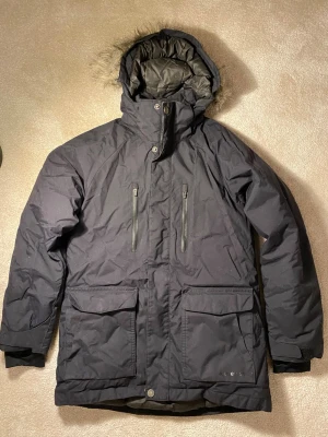 Svart parkasjacka med fuskpäls - Snygg svart parkasjacka med huva och fuskpälsdetalj. Jackan har två stora framfickor med lock, två bröstfickor med dragkedja och hög krage. Perfekt för kalla dagar och har en klassisk, rak passform. Man kan justera midjan med dragsko på insidan. 