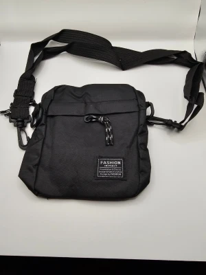 Svart crossbody-väska med patch Axelremsväska  - Snygg svart crossbody-väska i slitstarkt tygmaterial med justerbar axelrem och dekorativ patch framtill. Väskan har ett lock med snördetalj och praktisk karbinhake på sidan. Perfekt för att bära det viktigaste i vardagen.