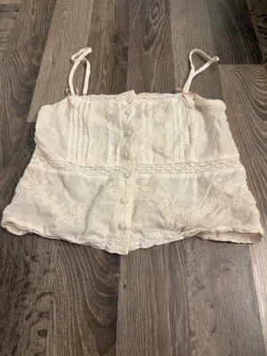 Vit spetsdetaljerad linnetopp - Söt vit linnetopp med tunna axelband, knappar framtill och fina spetsdetaljer. Toppen har dekorativa sömmar och broderier. Perfekt för varma dagar.