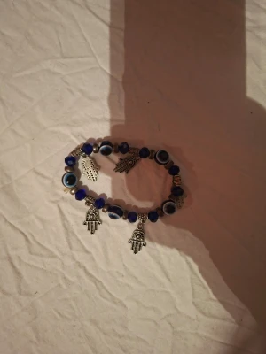 Blått armband med ögon och hamsa - Säljer ett elastiskt armband med blå och svarta pärlor, dekorerat med ögonpärlor och flera silverfärgade hamsa-händer som hängen. Armbandet har en bohemisk vibe och passar perfekt för dig som gillar unika detaljer och symbolik.