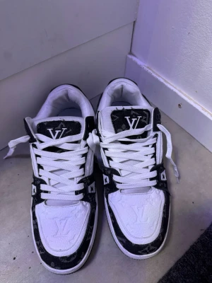Louis Vuitton svartvita sneakers - Säljer ett par snygga Louis Vuitton sneakers i svart och vitt med klassiskt LV-mönster på tå och sidor. Skorna har vita skosnören, låg profil och tydliga logodetaljer på plösen och sidan. Materialet är en mix av läder och textil.