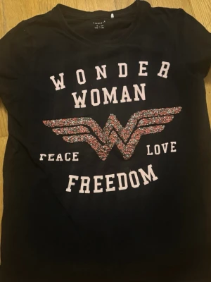Svart Wonder Woman t-shirt - Cool svart t-shirt från Name It med Wonder Woman-tryck i rosa och glitterdetaljer på bröstet. Texten 'Peace Love Freedom' och det ikoniska Wonder Woman-emblemet gör den extra unik. Perfekt för dig som vill sticka ut med superhjältestil.
