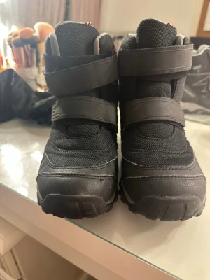 Svarta vinterkängor med kardborreband - Säljer ett par svarta vinterkängor med två breda kardborreband för enkel på- och avtagning. Kängorna har grov sula för bra grepp och är tillverkade i en kombination av syntetmaterial och textil. Insidan är fodrad för extra värme under kalla dagar.