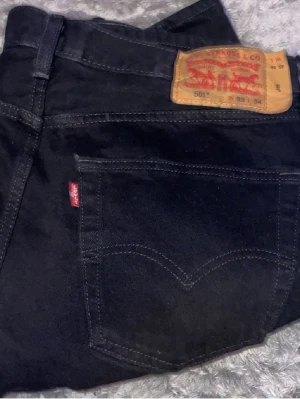 Svarta Levi's 501 jeans - Klassiska svarta Levi's 501 jeans med rak passform och femficksmodell. Jeansen har den ikoniska läderlappen bak i midjan och röd Levi's-tag på bakfickan. Tillverkade i slitstark bomull med tydliga sömmar och tidlös design.