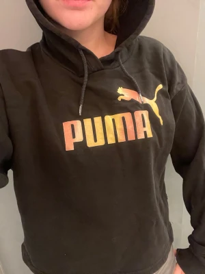 Svart cropped hoodie från Puma - Snygg svart hoodie från Puma med färgglatt puma tryck på bröstet. Hoodien har huva och långa ärmar. Perfekt för en chill och sportig look. Materialet är mjukt, känns luftigt och bekvämt, passar till både jeans och träningsbyxor. Lite kort vid mage, det är en typ av magtröja men det syns inte så mycket. Ej använt på länge. Ej bra pris,,,, skriv till mig 