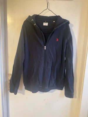 U.S. Polo Assn - Mörkblå hoodie från US Polo Assn med dragkedja framtill och två fickor. Klassisk röd logga broderad på bröstet. Snygg till ett par jeans. Storlek 14-15 vilket passar som xs. Nypris 649kr. 💕