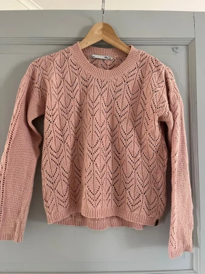 Rosa stickad tröja  - Söt ljusrosa stickad tröja från 157 med fint hålmönster och rund halsringning. Tröjan har långa ärmar och ribbade muddar vid ärmslut och nederkant. Perfekt för lager-på-lager och ger en mjuk, trendig look.