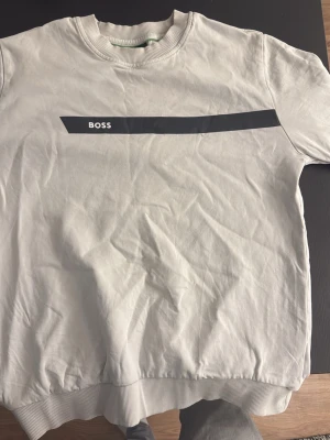 Vit tröja från BOSS - Vit tröja från BOSS med svart tryck över bröstet där det står 'BOSS'. Klassisk rund halsringning och ribbade muddar vid ärmslut och nederkant. Perfekt för en clean och stilren look.