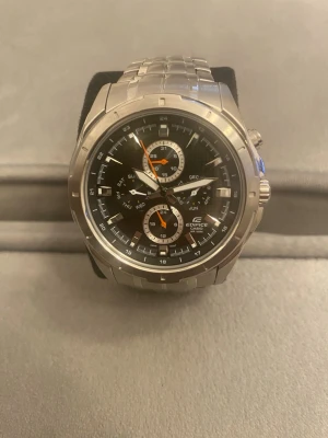 Casio Edifice silverfärgad klocka - Snygg Casio Edifice klocka i silver med länkarmband i metall. Urtavlan är svart med flera små urtavlor och detaljer i vitt och orange. Klockan har kronograf och datumvisning, samt robust design som passar till många stilar. Behöver byta batteri 