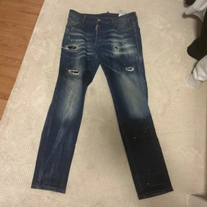 Blå slitna jeans med hål - Säljer ett par blå jeans med slitningar, hål och färgstänk för en trendig och avslappnad look. Jeansen har raka ben, fem fickor och knappgylf. Perfekta för dig som gillar streetwear och en edgy stil.