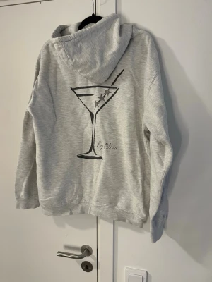 Ljusgrå hoodie med martiniglas-tryck - Ljusgrå hoodie med ett stort tryck av ett martiniglas med tre oliver på ryggen. Mjuk och skön och perfekt för myskvällar eller kyligare dagar