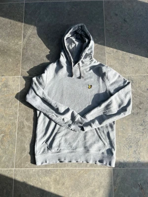 Grå hoodie från Lyle & Scott - Snygg grå hoodie från Lyle & Scott med klassisk gul logga på bröstet. Hoodien har justerbar dragsko i huvan och en stor magficka framtill. Tillverkad i mjukt sweatshirtmaterial som är perfekt för chill dagar. Ribbade muddar vid ärmslut och nederkant. Super fint skick på den och märks knappt att den använts. Hör av er om ni har frågor!