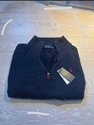 Mörkblå halvzip från Polo Ralph Lauren - Klassisk mörkblå stickad tröja från Polo Ralph Lauren med halvzip och ribbad krage. Tröjan har det ikoniska röda Polo-logot på bröstet och är tillverkad i mjukt material som ger en clean och stilren look.Box ingår inte samma med tag.Men man kan betala extra om man vill ha med prislapen och tagen